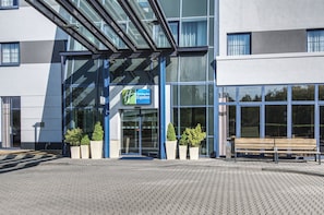 Un bâtiment de l’hôtel Holiday Inn Express avec une entrée en verre, des plantes en pot et un banc à l’extérieur.