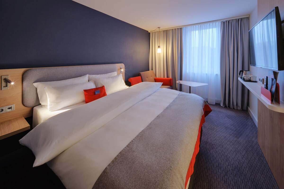Une chambre d’hôtel avec un grand lit, un bureau, une télévision et une fenêtre avec des rideaux.