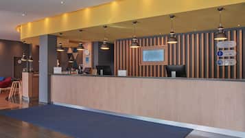 A hotel reception area with a wooden reception desk, multiple pendant lights, a clock, and a sign indicating the General Manager's office.