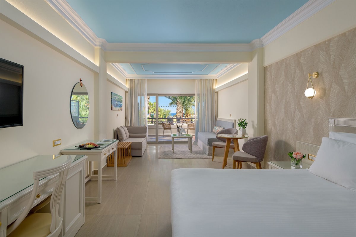 Ein Hotelzimmer mit Bett, Schreibtisch, Sessel, Sofa und einem Balkon mit Blick auf Palmen.