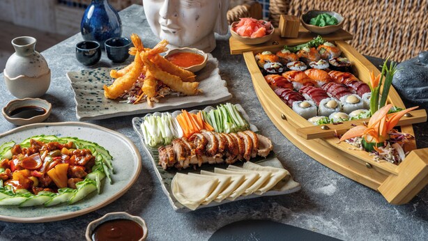 Une variété de plats, notamment des sushis, des beignets japonais et un plat de viande, présentés sur une table agrémentée d’une sculpture de tête de Bouddha et de petits bols de sauce.