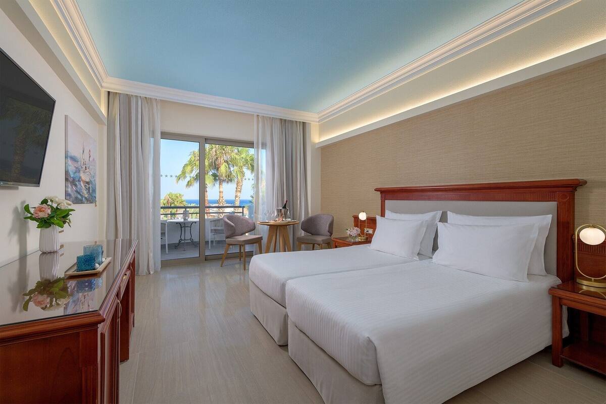 Ein Hotelzimmer mit zwei Betten, einem Balkon mit Blick auf Palmen und Meer, einem Schreibtisch mit Stuhl und einem an der Wand montierten Fernseher.