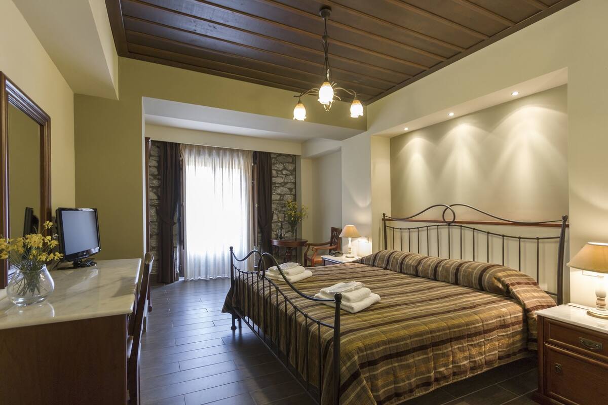 A bedroom with a bed, bedside table, lamp, and a desk with a computer monitor and vase of flowers.