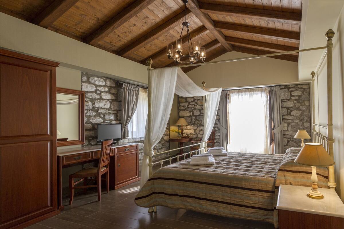 A bedroom with a stone wall, a wooden desk, a bed with a canopy, and a chandelier.