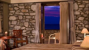 A bedroom with a stone wall, a bed, a chair, a table with a candle, and a view of the sea at dusk. - Itilo Traditional Hotel (East Mani)