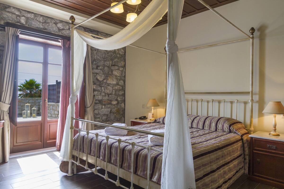 A four-poster bed with a stone wall and a view through a glass door.