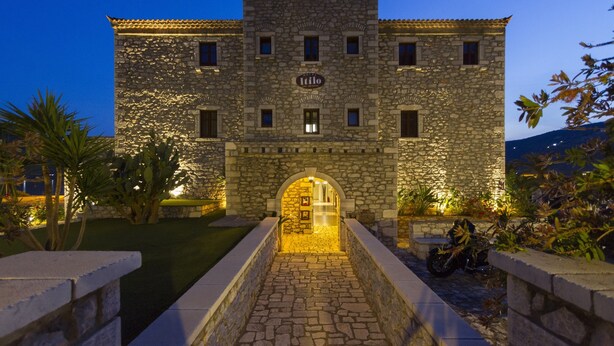 A stone building with a sign reading "Italo" at the entrance, surrounded by greenery and a paved pathway. - Itilo Traditional Hotel (East Mani)