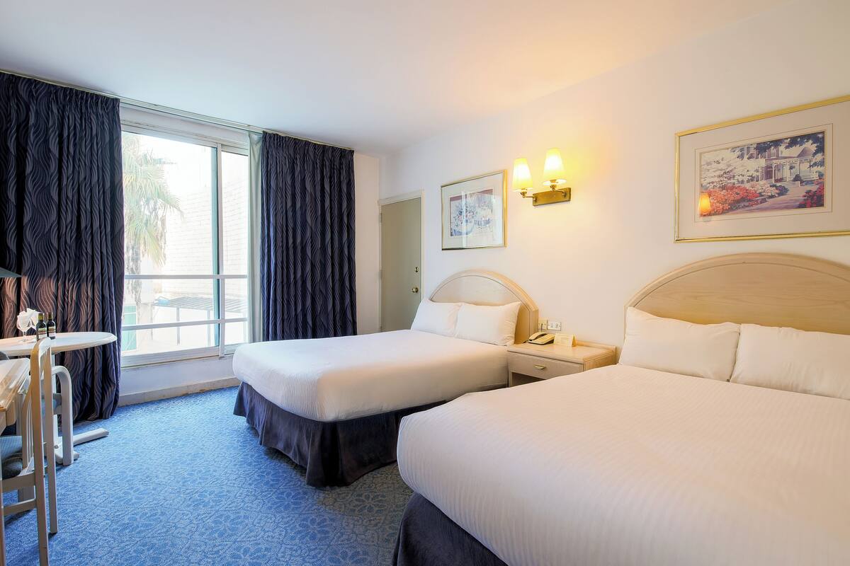 A hotel room with two beds, a desk, and a chair. There is a window with curtains, a wall-mounted lamp, and a framed picture.