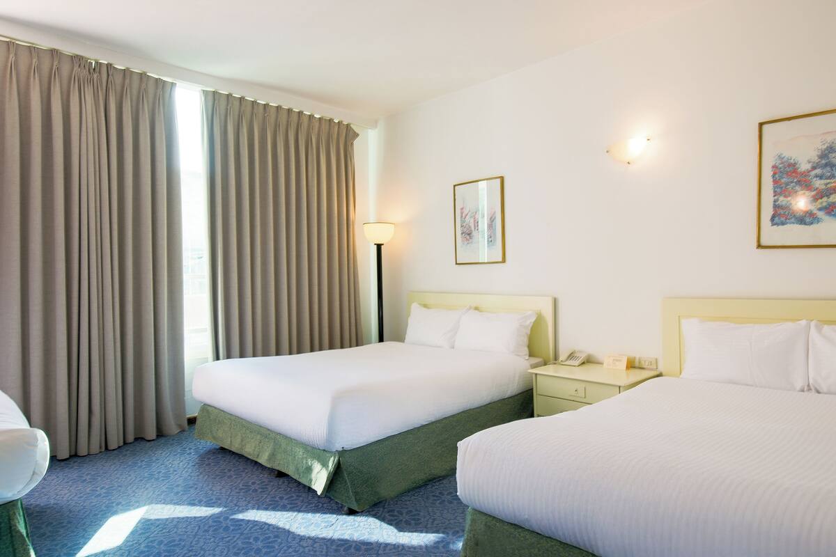 A hotel room with two beds, a bedside table with a lamp, a framed picture on the wall, and a window with curtains.