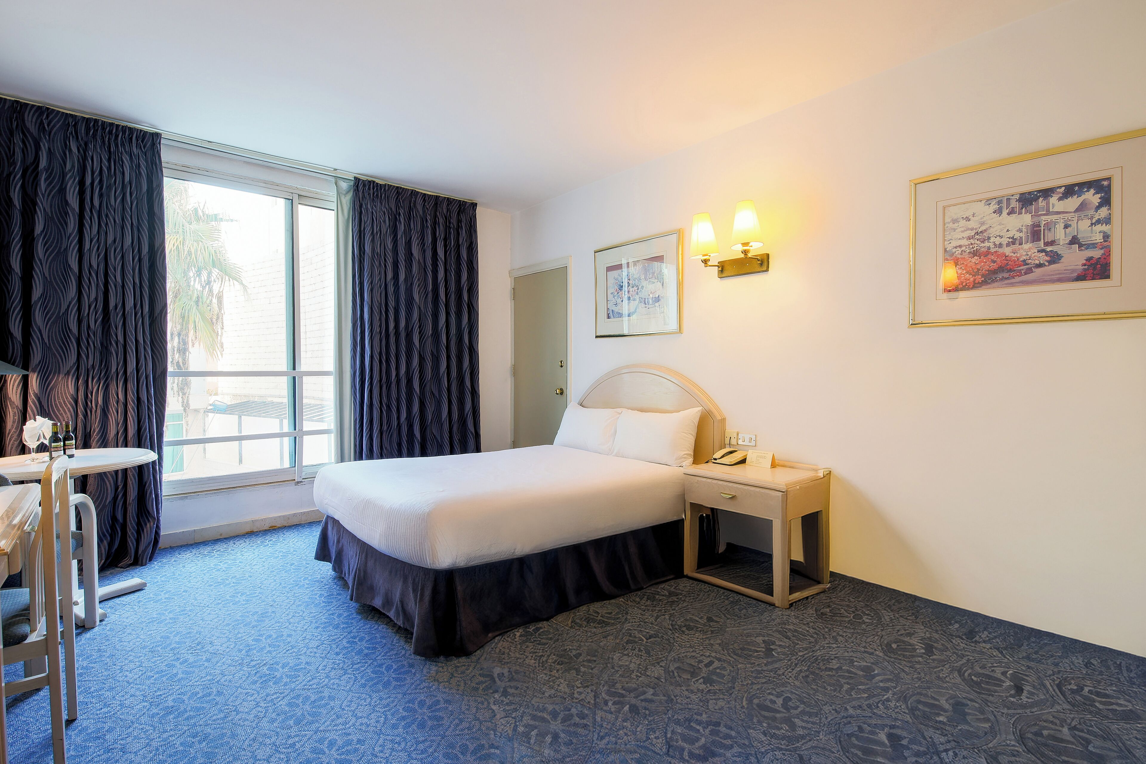 Quarto de hotel com cama, mesa de cabeceira, abajur e janela com cortinas.