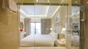 A modern hotel room with a glass-enclosed shower, a large bed, a bedside table with a lamp, and a view of the city through the window. - Petra Palace Hotel (Wadi Musa)