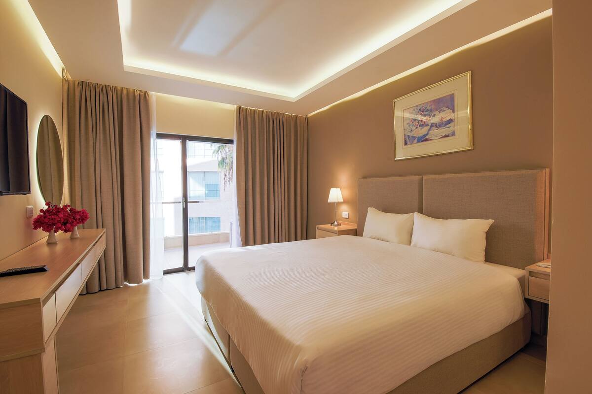 A hotel room with a large bed, a bedside table with a lamp, a flat-screen TV, and a painting on the wall.