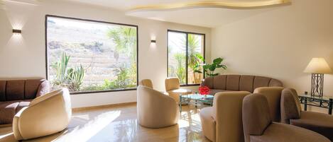 A modern living room with a beige sofa, two armchairs, a glass coffee table, and a lamp. Large windows provide natural light and a view of greenery outside.