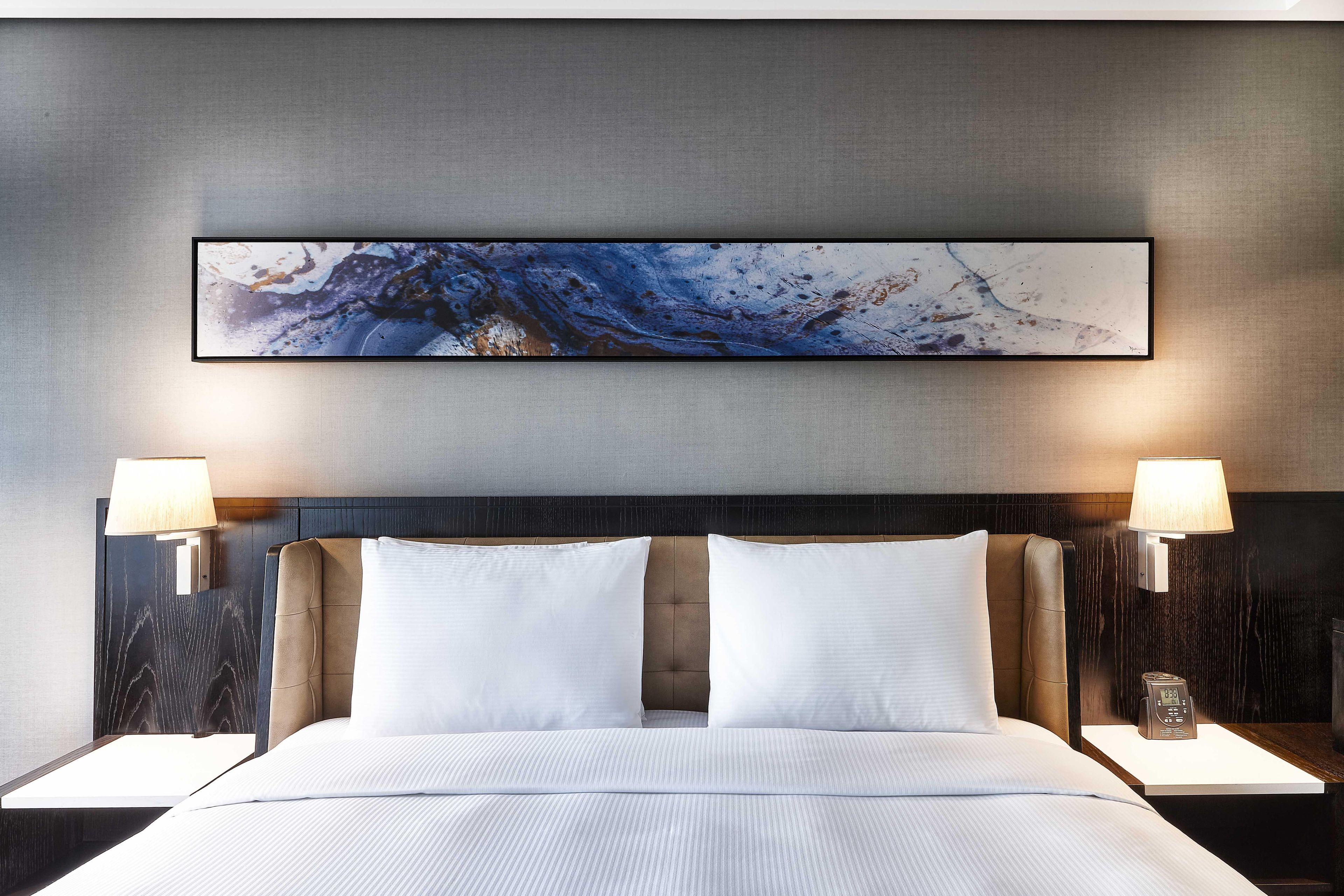 A hotel room with a large bed, two bedside tables, a wall-mounted abstract painting, and two wall-mounted lamps.