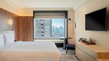 Quarto de hotel com uma cama grande, uma escrivaninha, uma cadeira, uma televisão e uma janela com vista para a cidade.