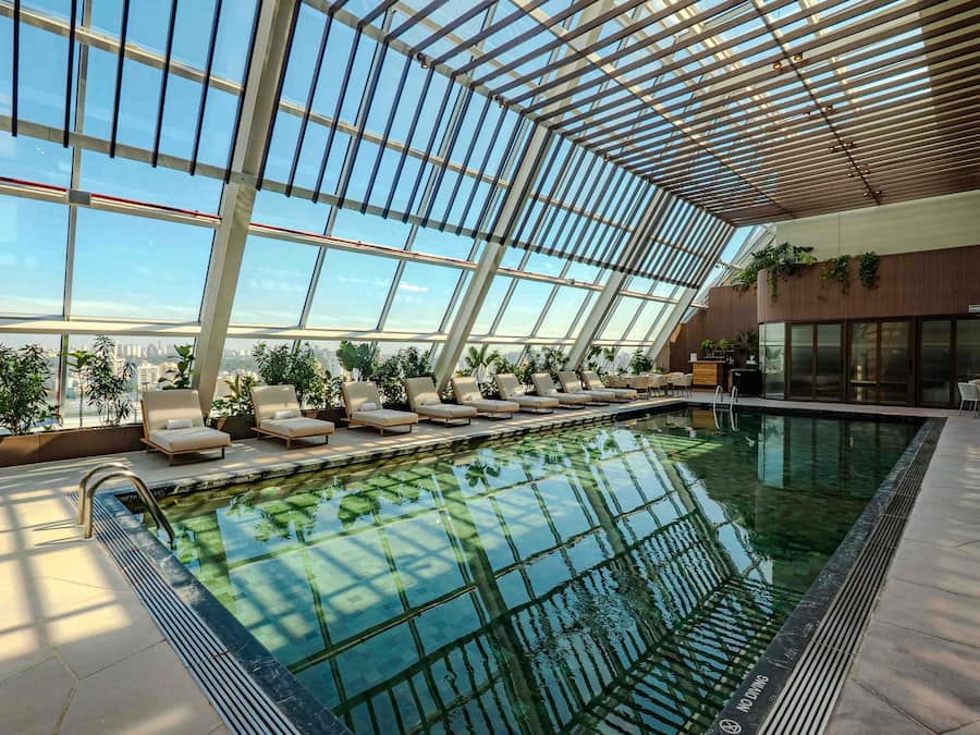 Piscina interna com teto de vidro, espreguiçadeiras e vista da cidade.