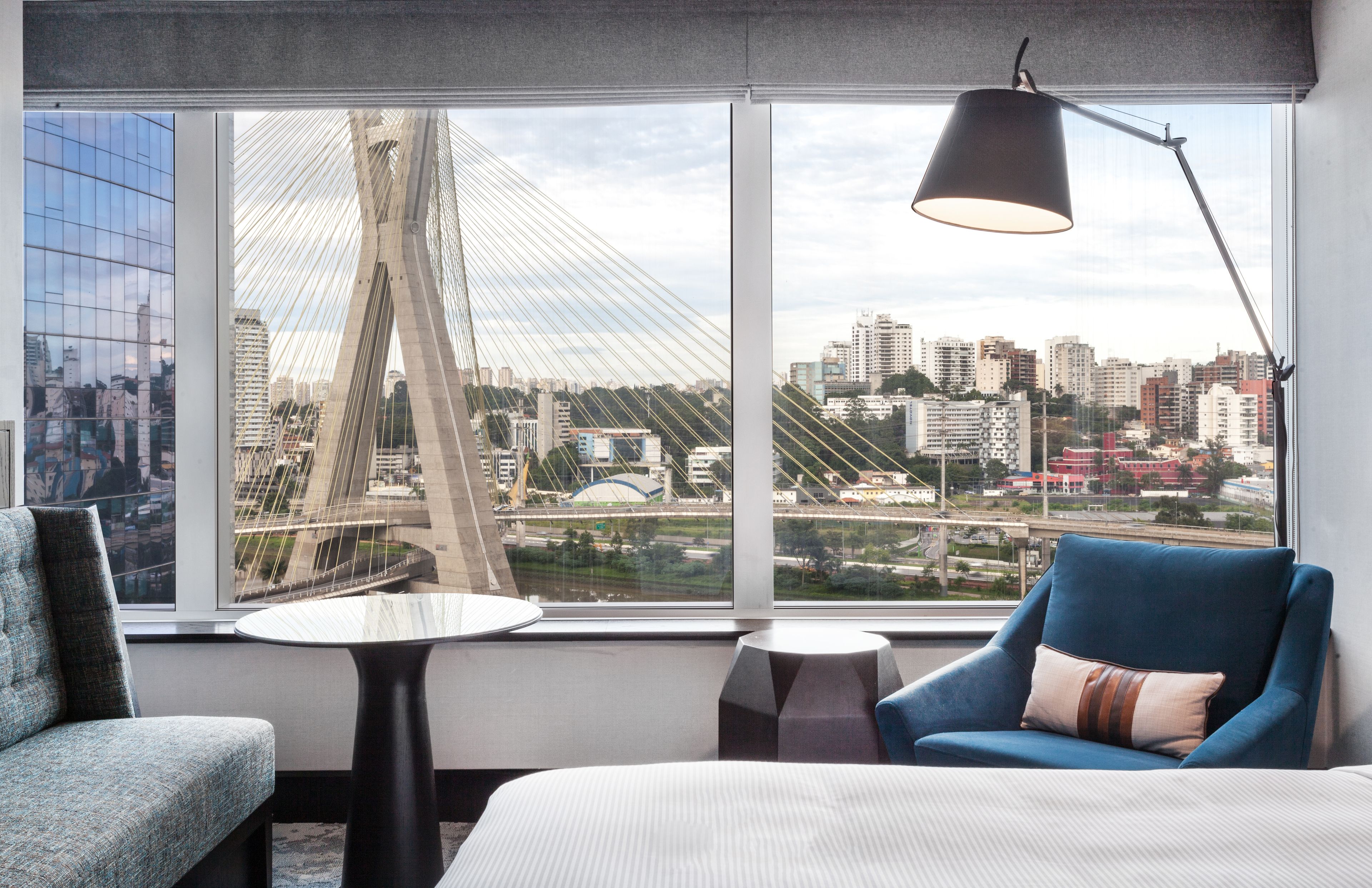 Une chambre d’hôtel moderne dotée d’une grande fenêtre offrant une vue sur les gratte-ciel de la ville et sur un pont. On y trouve un fauteuil bleu, une petite table ronde et une lampe.