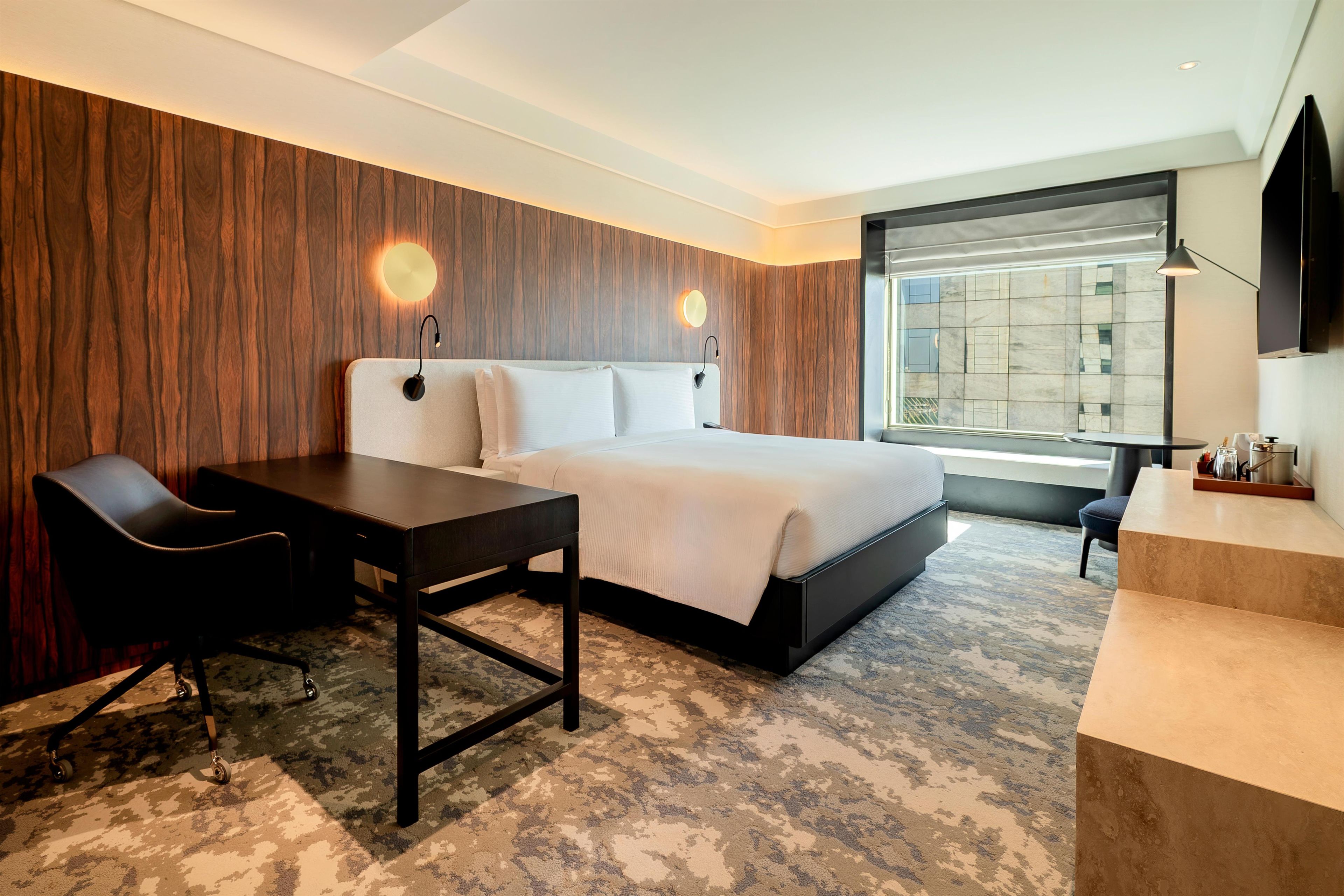Ein modernes Hotelzimmer mit einem großen Bett, einem Schreibtisch, einem Stuhl und Blick auf die Stadt durch ein großes Fenster.