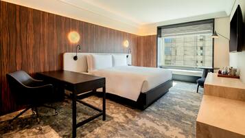 Ein modernes Hotelzimmer mit einem großen Bett, einem Schreibtisch, einem Stuhl und Blick auf die Stadt durch ein großes Fenster.