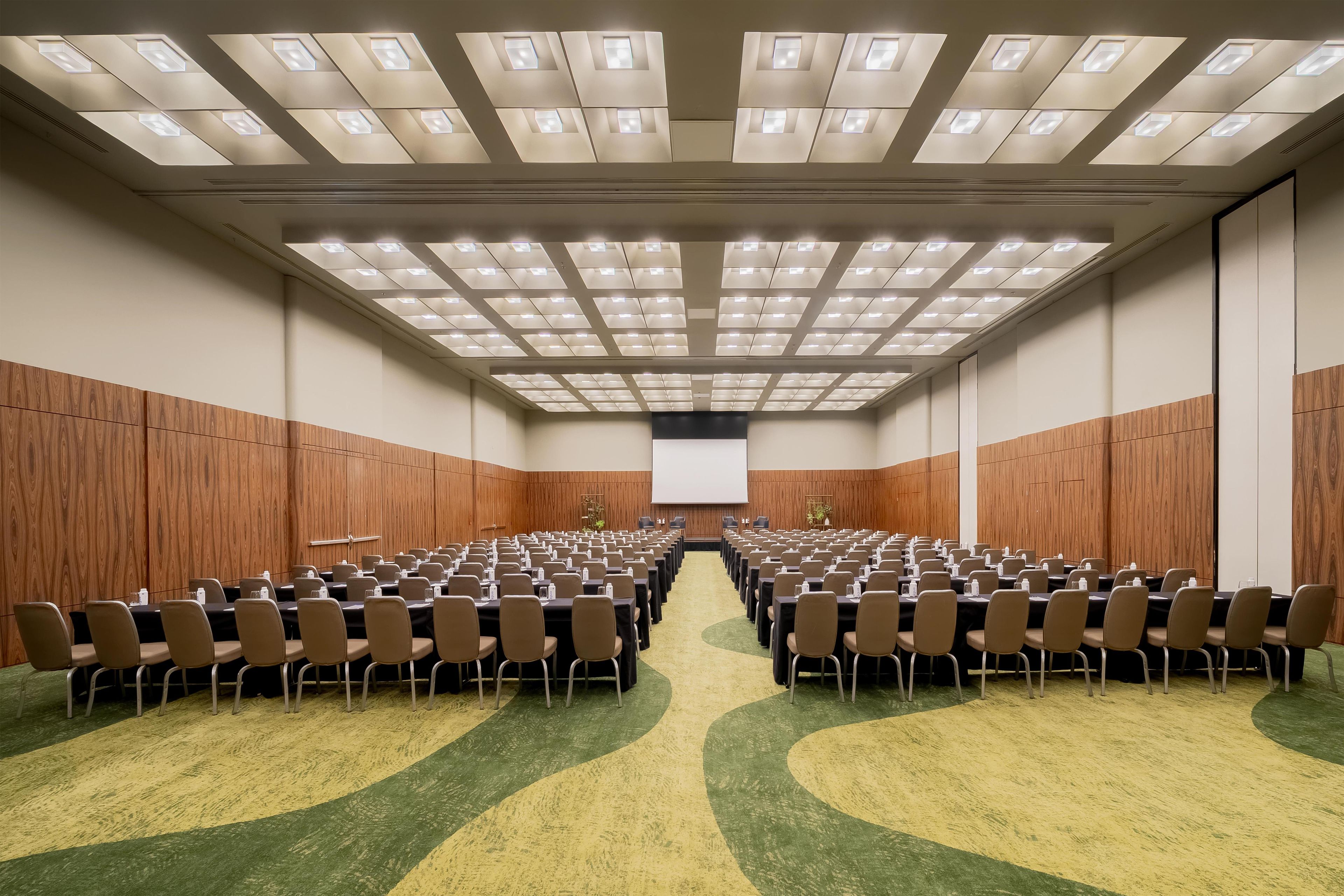 Een congreszaal met rijen tafels en stoelen, een projectiescherm en een wand van houten panelen.