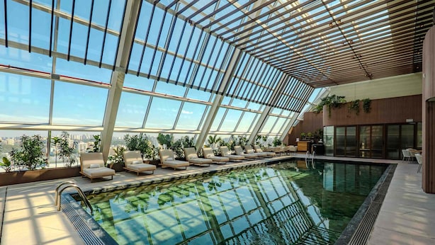 An indoor swimming pool with a glass ceiling, lounge chairs, and a view of the city.