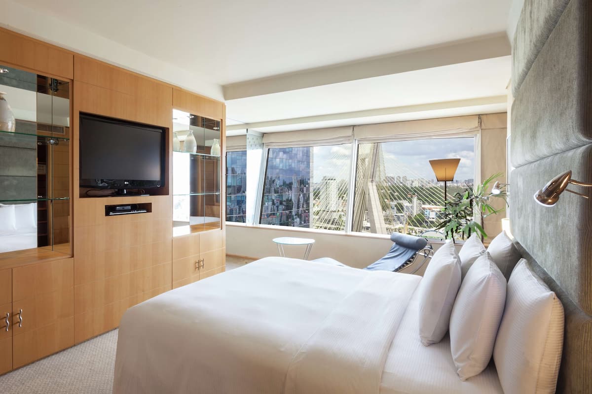 Una habitación de hotel moderna con una cama grande, un televisor, una mesita de vidrio y vistas a un paisaje urbano a través de amplias ventanas.