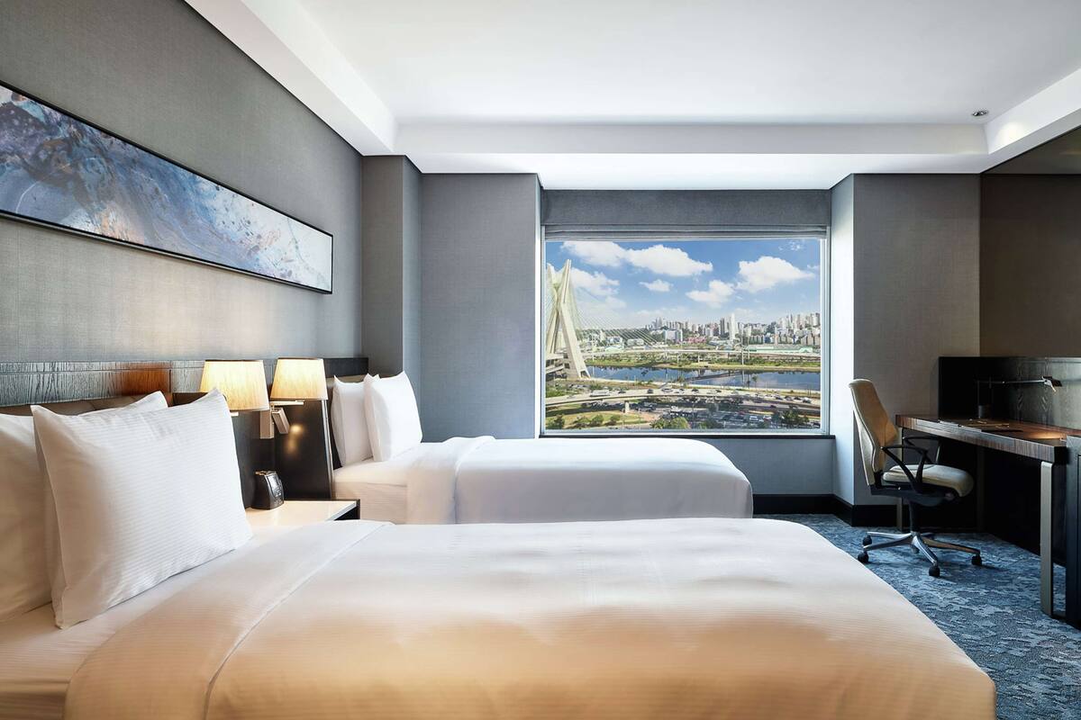 Habitación de hotel con dos camas, un escritorio, una silla y un amplio ventanal con vista a la ciudad.