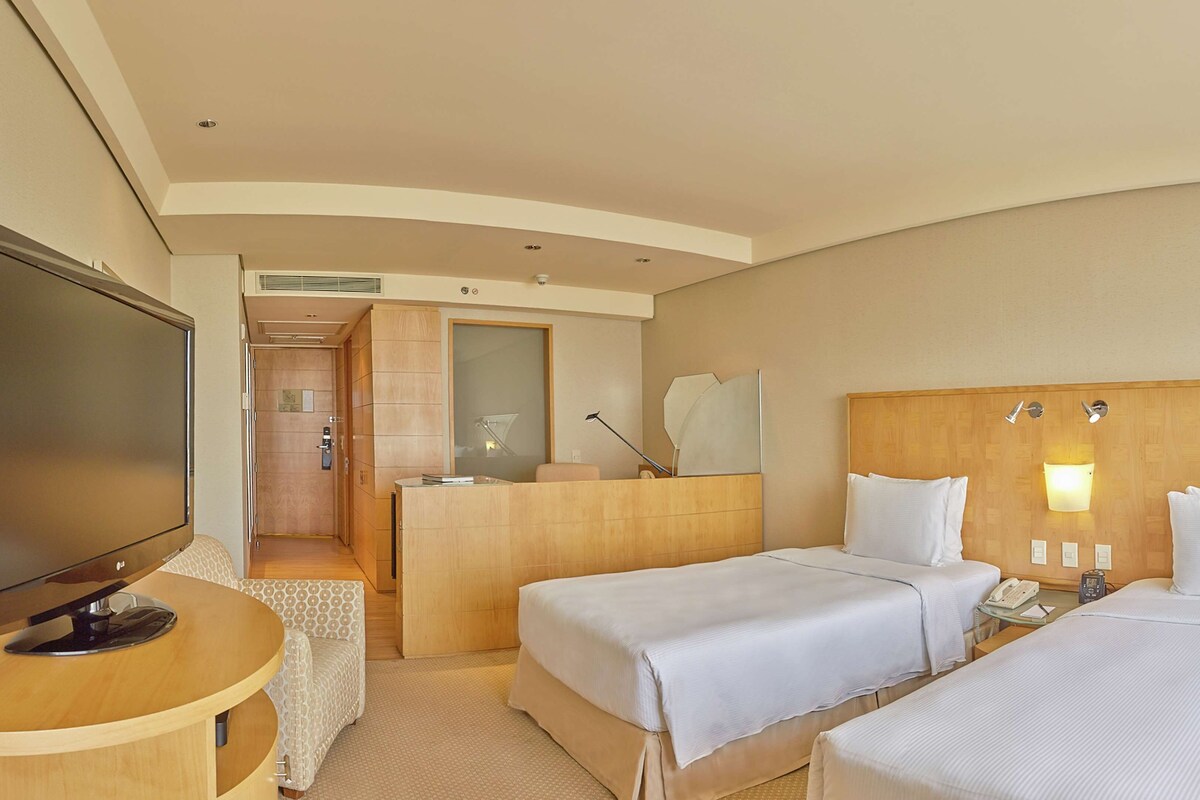 A hotel room with two beds, a TV, a desk, and a bathroom visible through an open door.