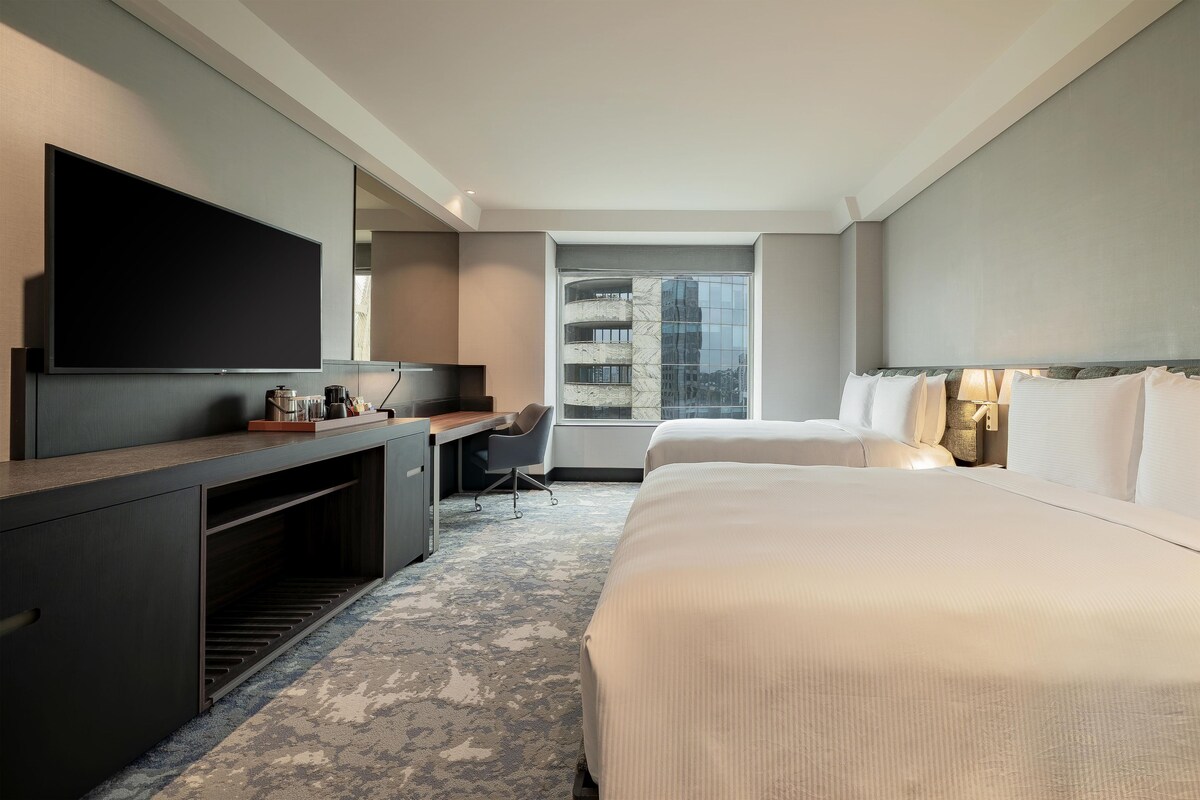 Une chambre d’hôtel avec un grand lit, un bureau doté d’un téléviseur à écran plat, une chaise et une vue sur le paysage urbain par une grande fenêtre.