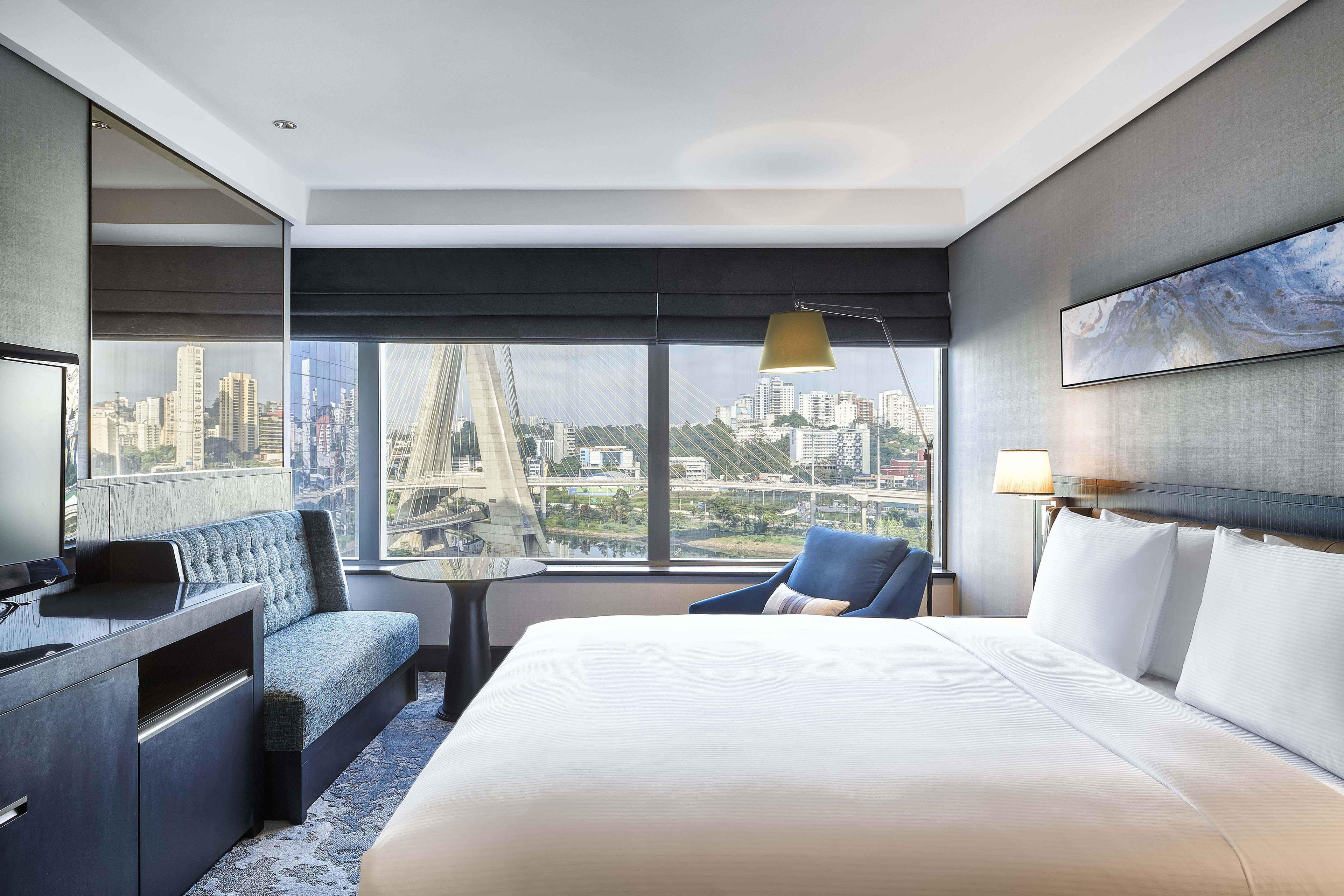 Una habitación de hotel con cama, escritorio, silla, televisor y un amplio ventanal con vista a la ciudad.