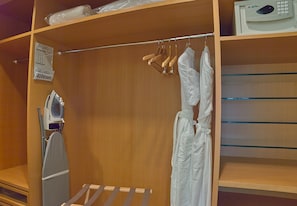 Een houten kast met hangende kleding, een wit strijkplank en een magnetron op een plank.