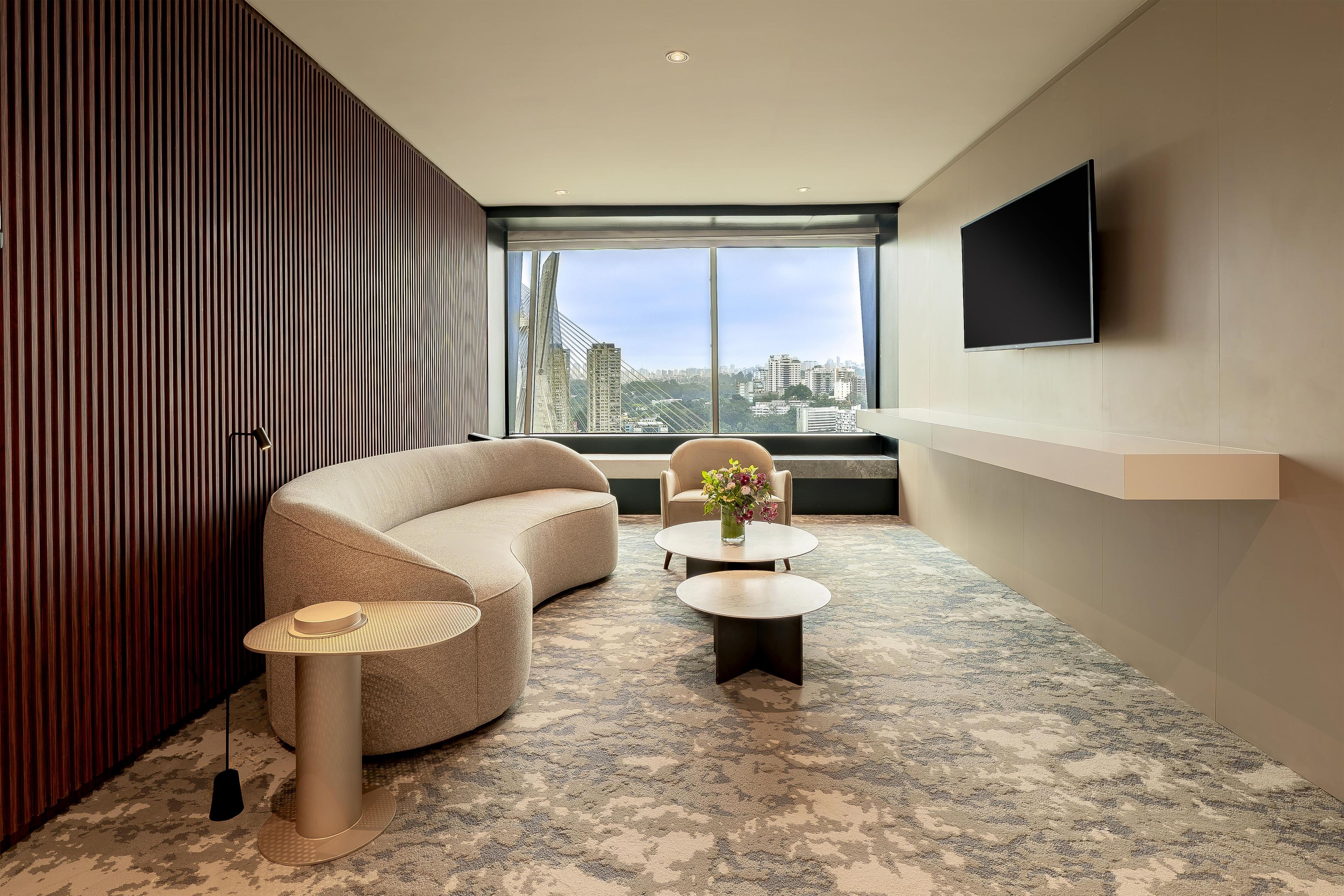 A modern hotel room with a large window, a sofa, two side tables, and a flat-screen TV mounted on the wall.