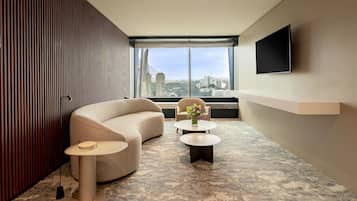 A modern hotel room with a large window, a sofa, two side tables, and a flat-screen TV mounted on the wall.