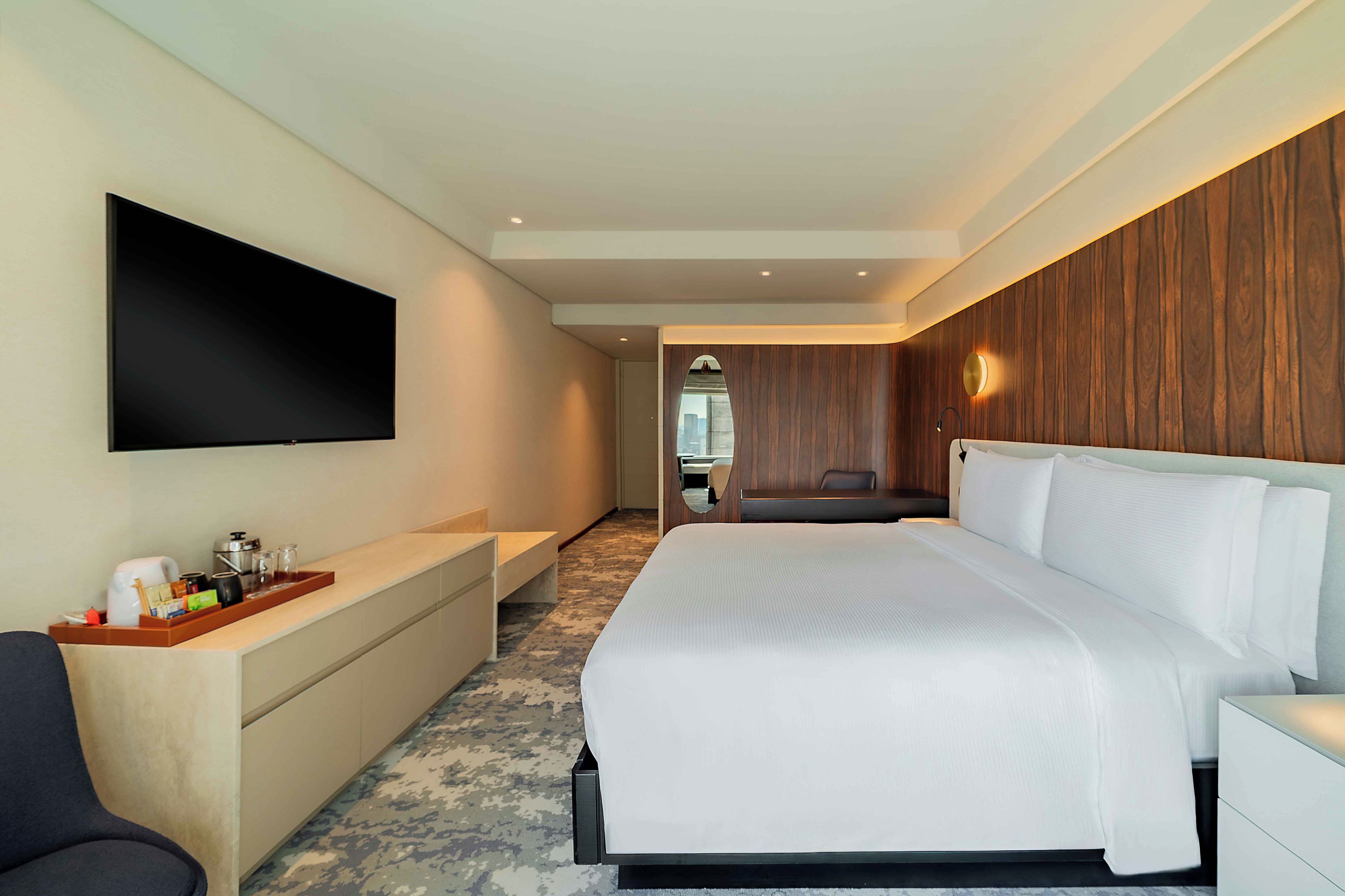 A modern hotel room with a large bed, a flat-screen TV mounted on the wall, a seating area with a view, and a wooden accent wall.