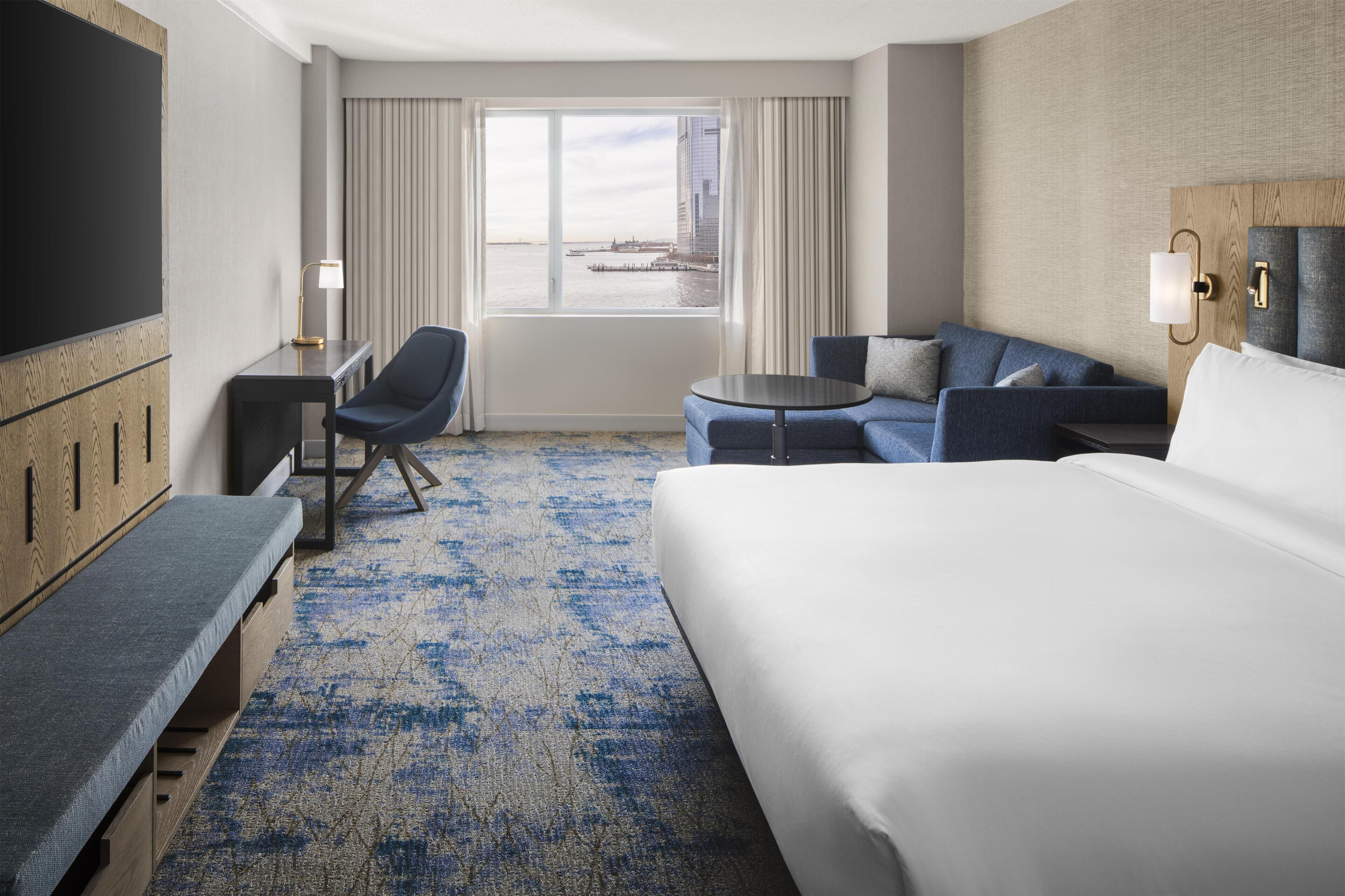 Une chambre d’hôtel avec un grand lit, un canapé, un bureau et une chaise. Il y a une fenêtre donnant sur la ville et sur un plan d’eau.