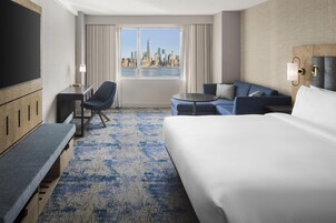 A hotel room with a large bed, a seating area with a sofa and armchairs, a desk with a chair, a TV, and a view of a city skyline through the window.