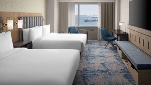 Ein Hotelzimmer mit zwei Betten, einem Schreibtisch, einem Sessel, einem Fernseher und Blick auf das Wasser.