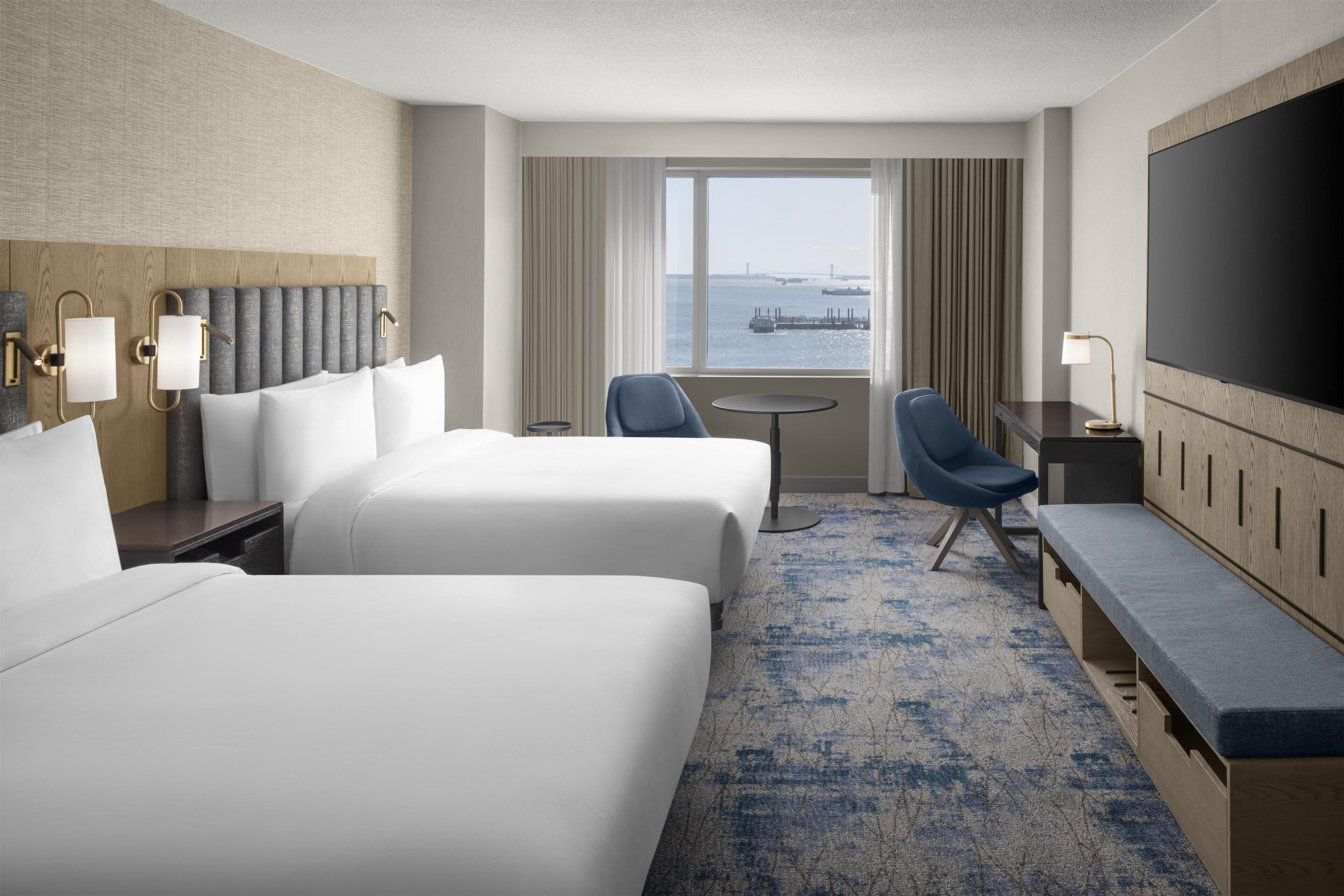 A hotel room with two beds, a desk, a chair, a TV, and a view of the water.