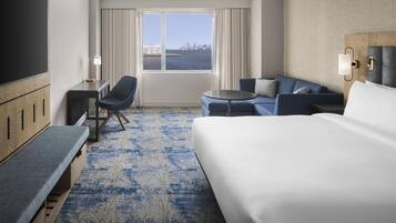 Quarto de hotel com uma cama grande, um sofá, uma escrivaninha com cadeira, televisão e janela com vista para a cidade.