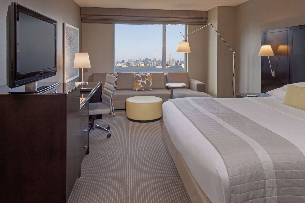 A hotel room with a bed, a desk with a TV, a sofa, a round table, and a city view.