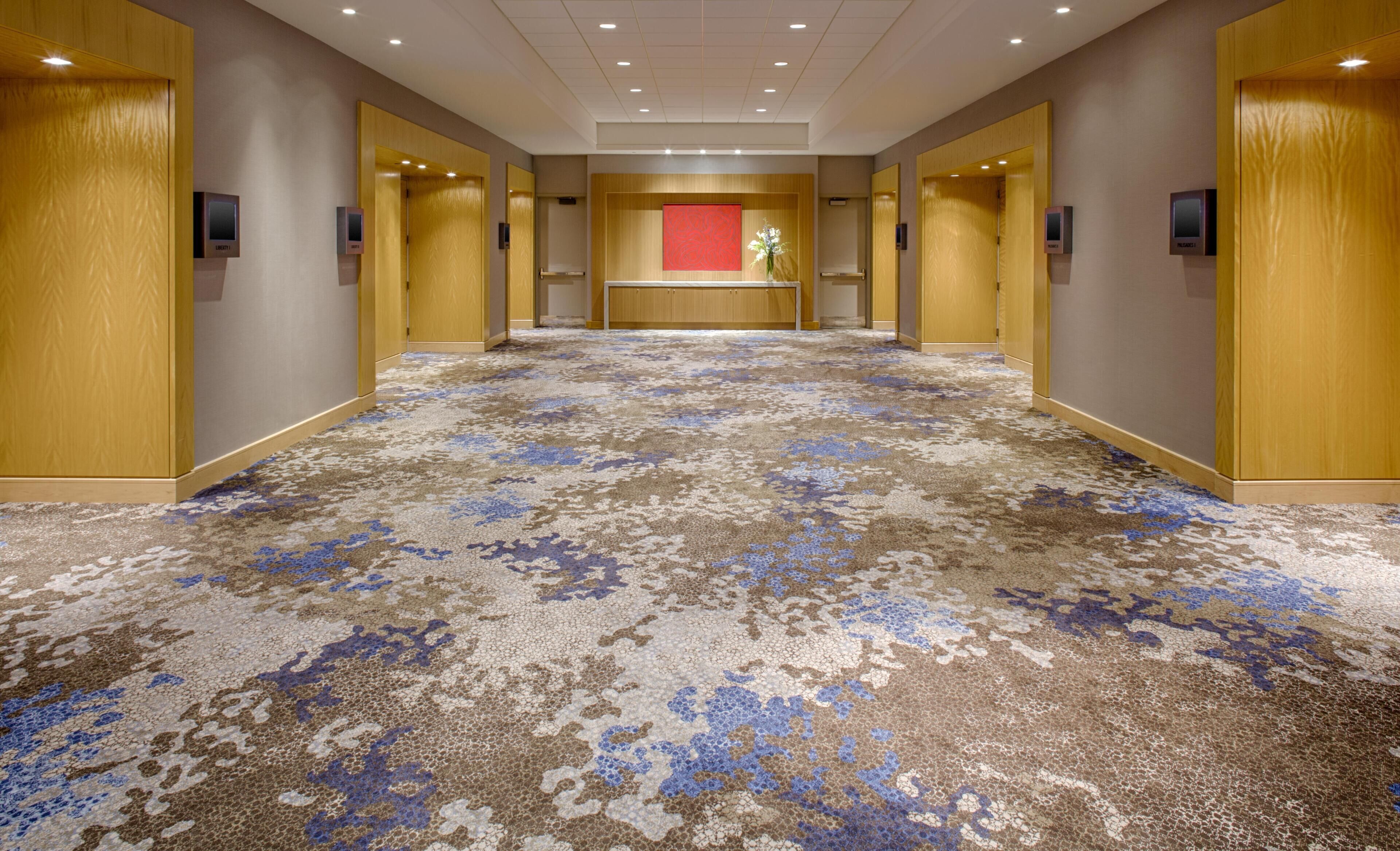 Un pasillo de hotel con alfombra estampada, puertas de madera y una recepción con pared de acento roja.