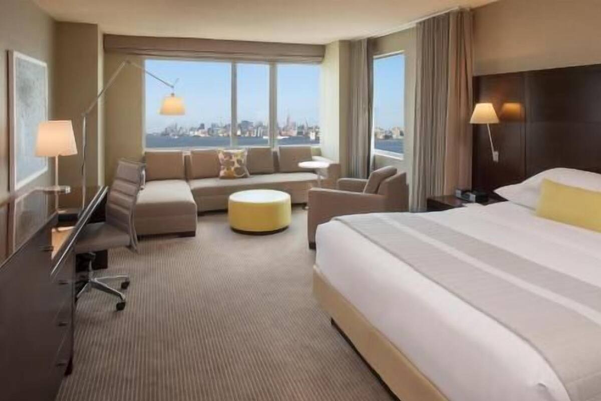 A hotel room with a large bed, a sofa, a small table, and a chair. There are two windows with a city view.