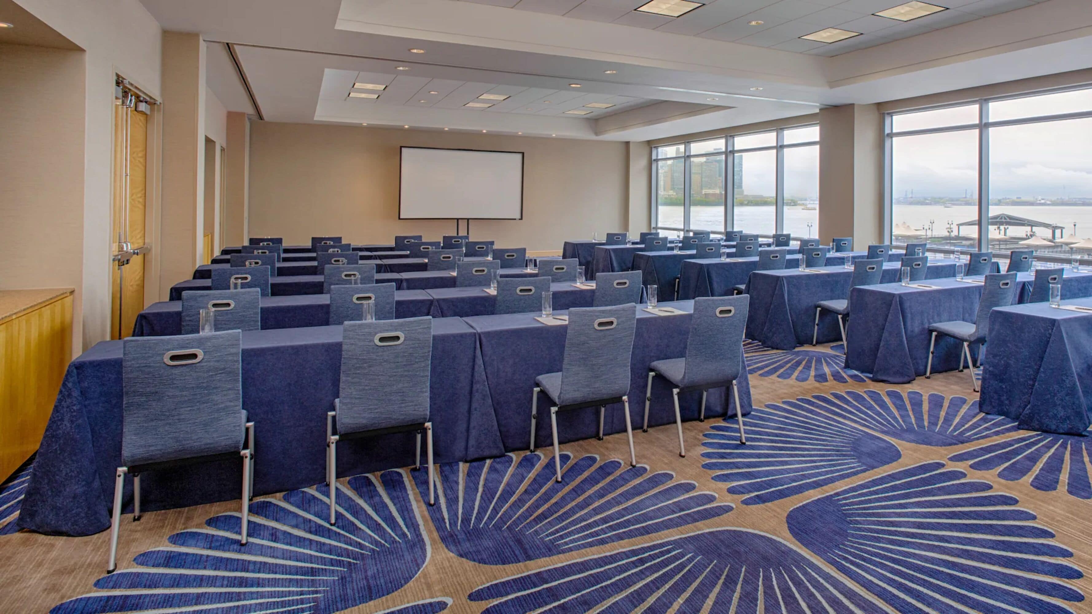 A conference room with rows of tables and chairs, large windows with a view of a cityscape, and a projector screen on the wall.