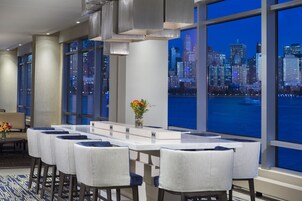 A modern hotel room with a large window offering a city view. There is a dining area with a white table and blue upholstered chairs.