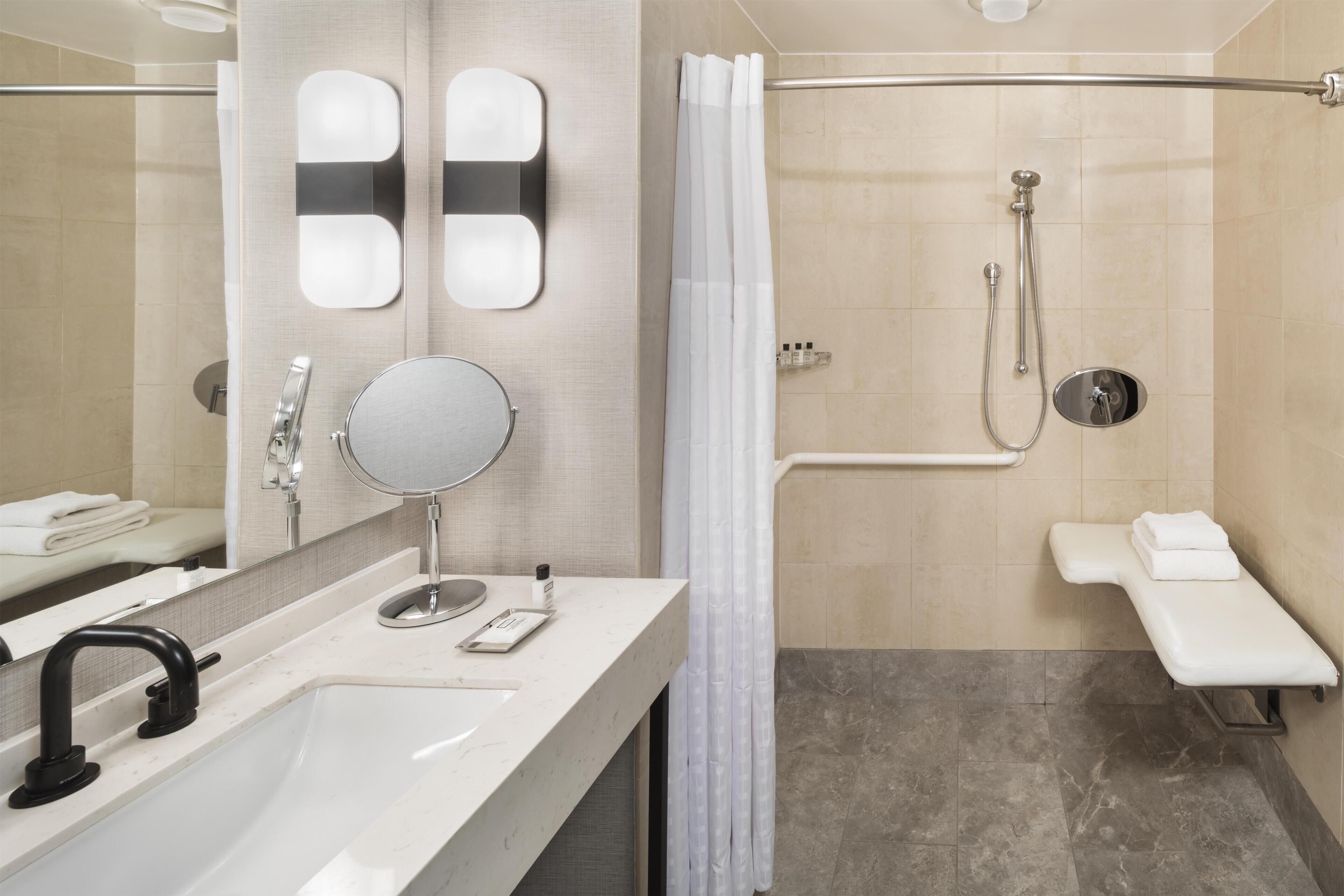A modern bathroom with a sink, mirror, shower, and a bench.