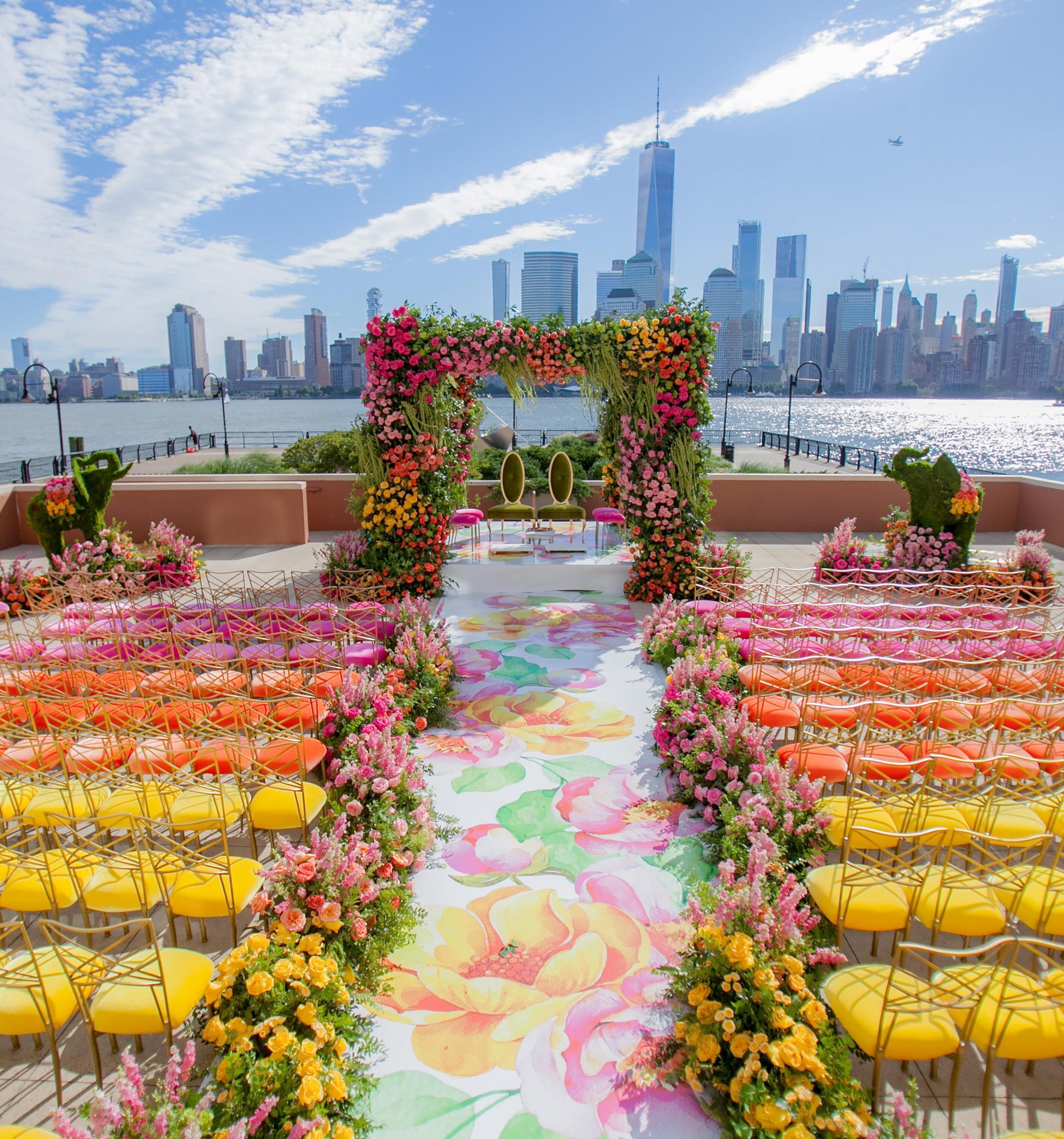 Un événement en plein air, avec une arche décorée de fleurs, des rangées de chaises et, en arrière-plan, la silhouette de la ville.