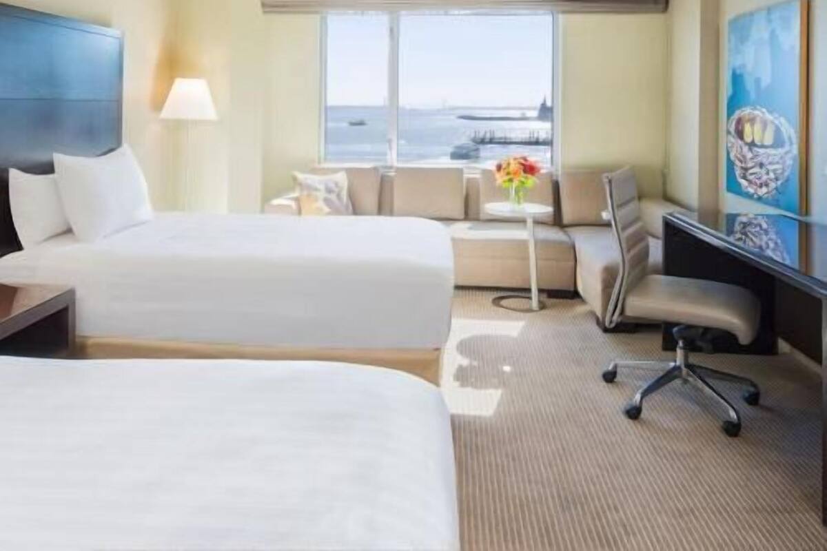 A hotel room with two beds, a desk, a chair, a window with a view, and a painting on the wall.