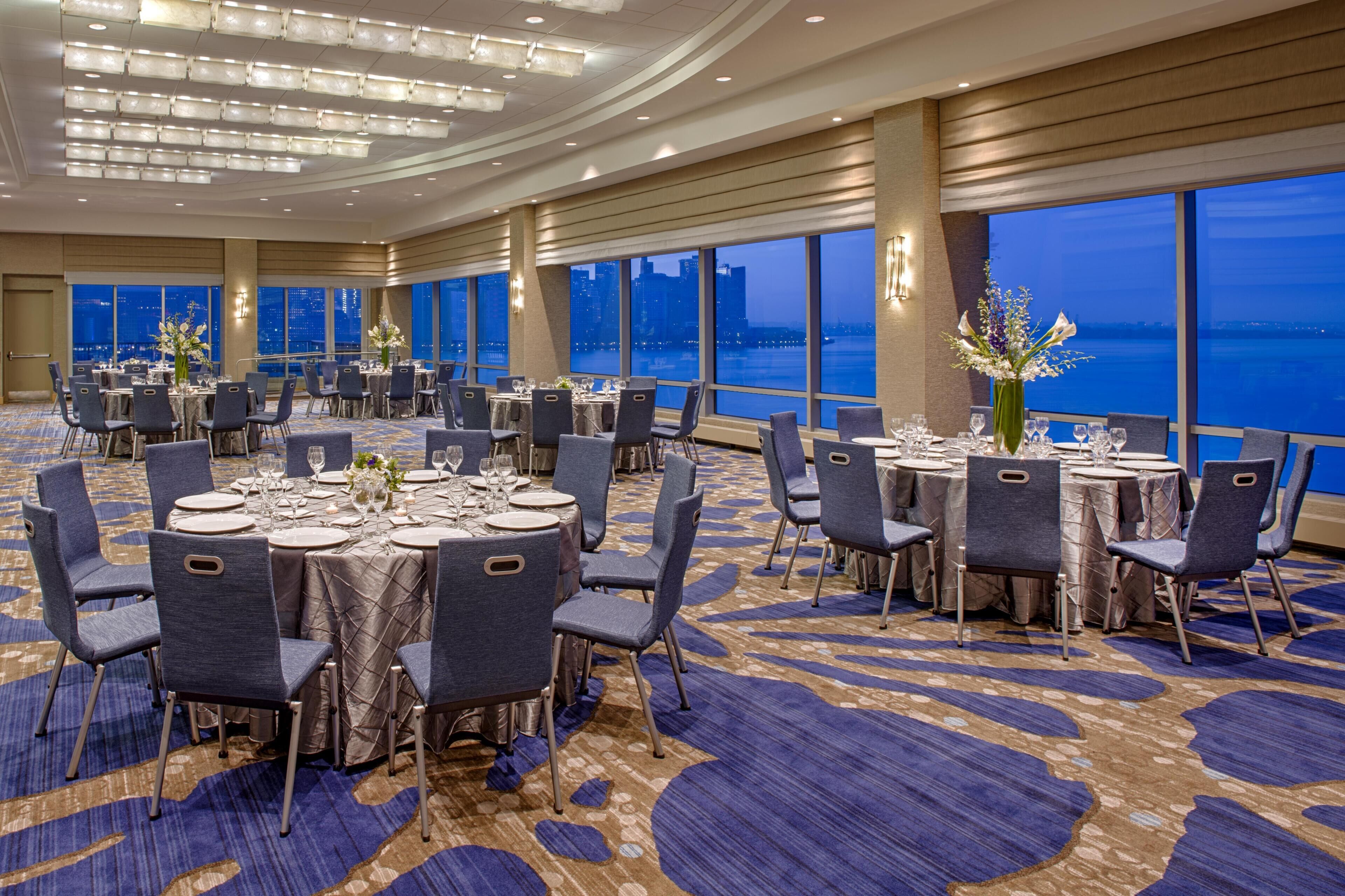 Uma sala de banquetes com mesas redondas, cadeiras e vista para a cidade.