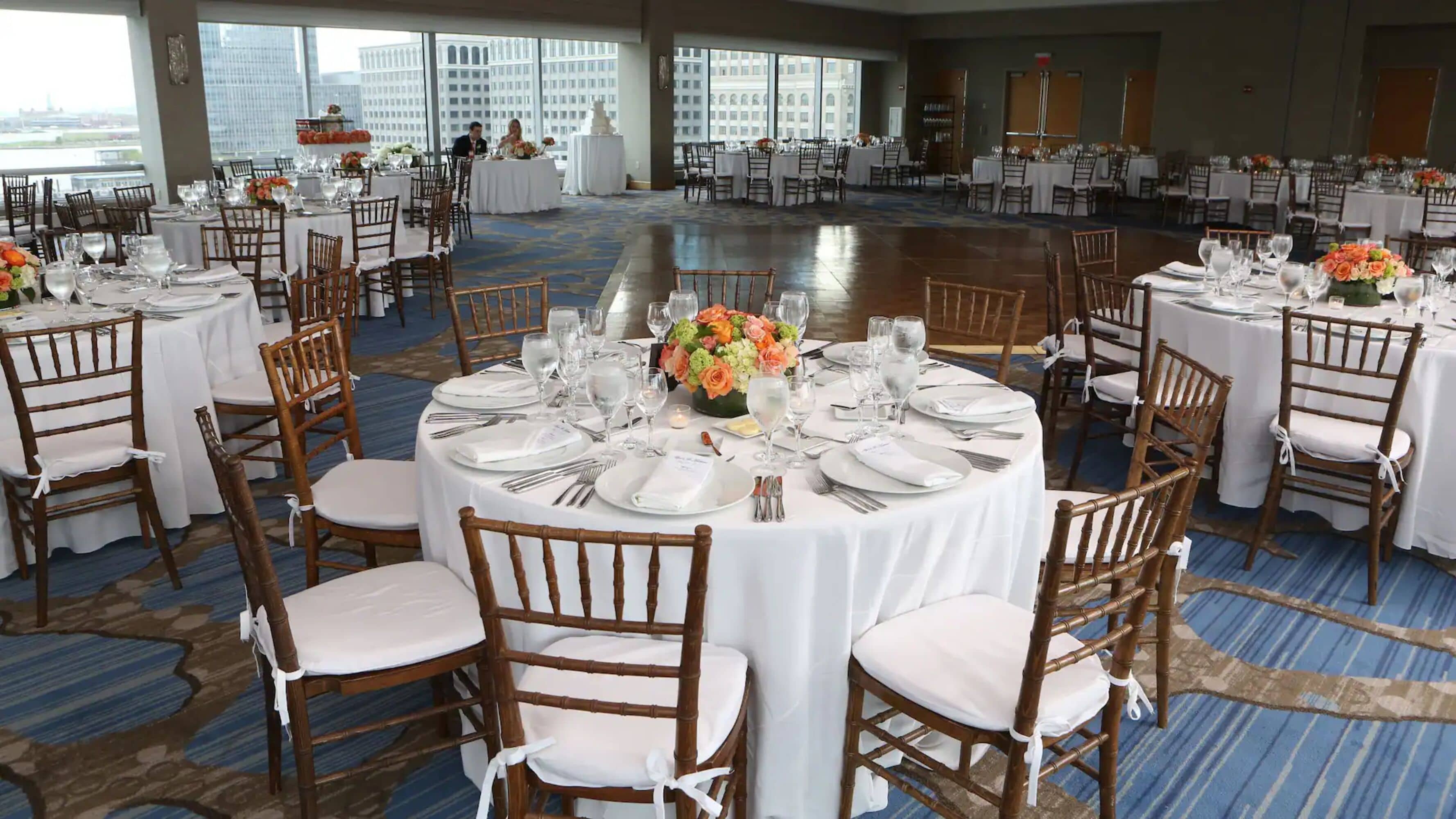 Un salón de banquetes con mesas redondas dispuestas para un evento formal, con manteles blancos, centros de mesa florales y sillas de madera.