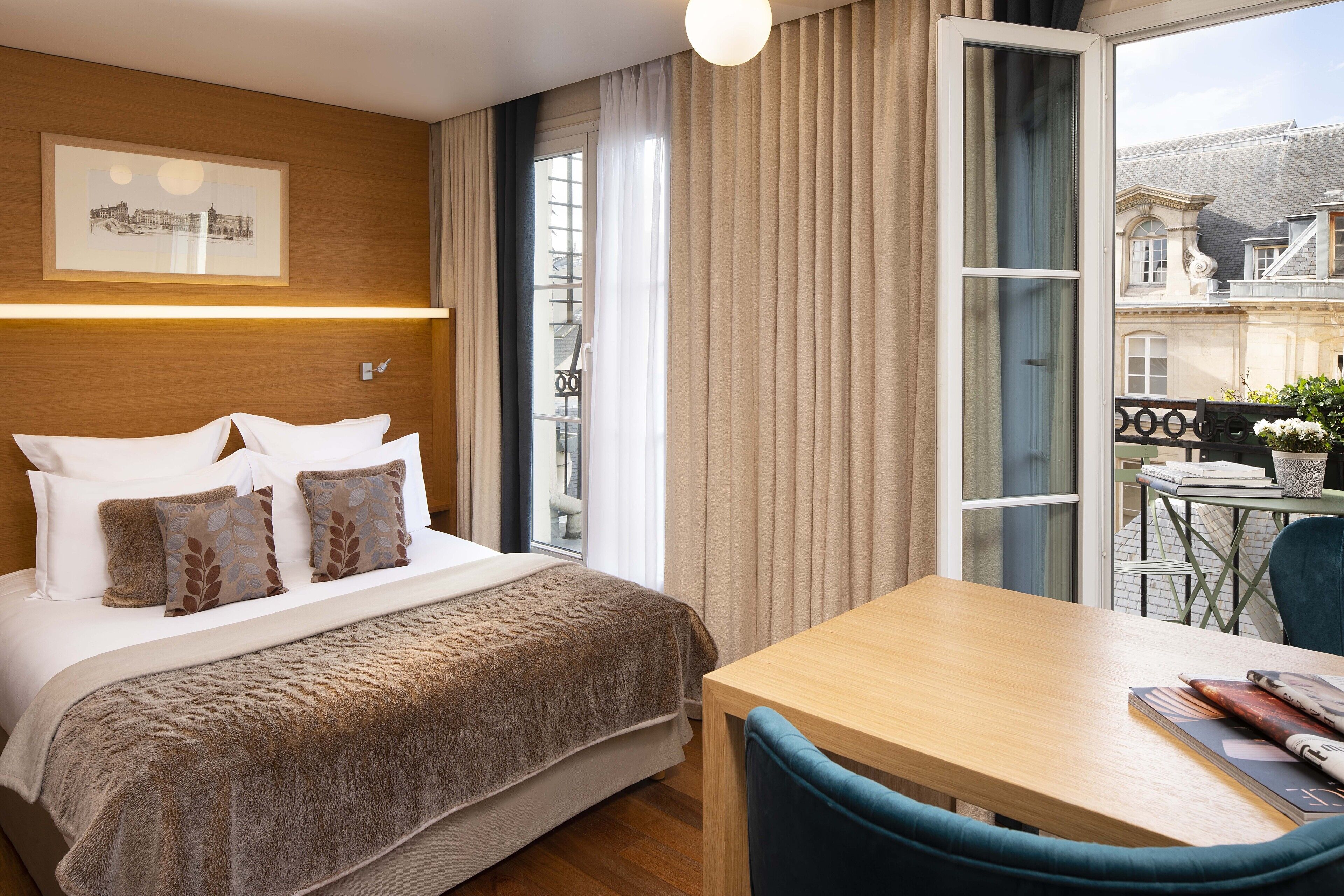Quarto de hotel com cama, cabeceira de madeira, mesa com cadeiras e vista para prédios pela janela.